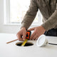 Hand met rolmaat meet het ronde zaaggat voor een inbouwspot in een strak wit plafond, met spotarmatuur en gereedschap op de achtergrond.